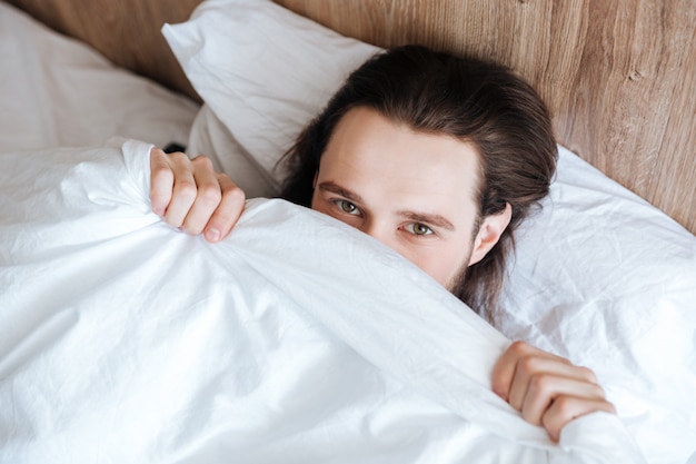 Przystojny mężczyzna ukrywa twarz pod białą narzutą w łóżku