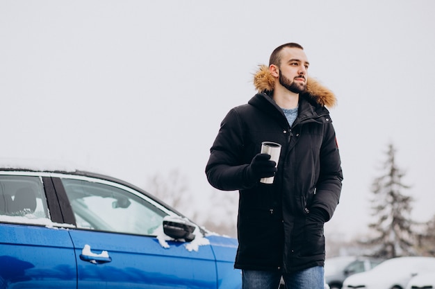 Przystojny mężczyzna w ciepłej kurtce stojący samochodem pokrytym śniegiem i picia kawy