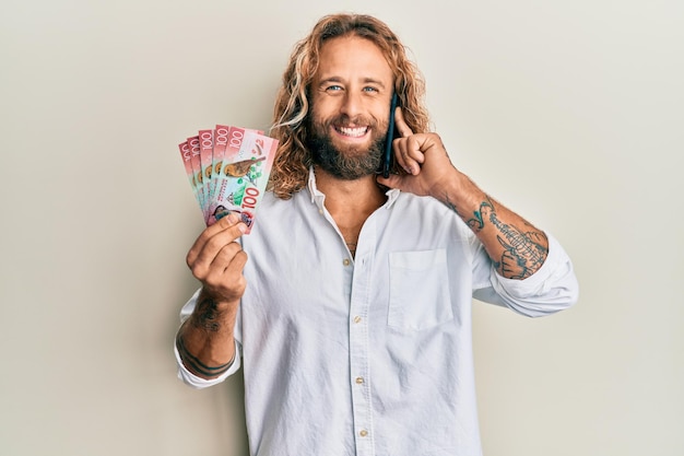 Przystojny mężczyzna z brodą i długimi włosami rozmawia przez telefon, trzymając 100 nowozelandzkich dolarów, uśmiechając się i śmiejąc się głośno, bo zabawny szalony żart.