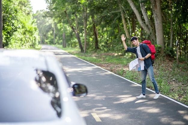 Bezpłatne zdjęcie przystojny mężczyzna z plecakiem i trzyma papierową mapę w ręku, podnosi rękę do autostopu samochodem na poboczu drogi, koncepcja autostopu