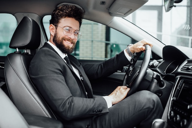 Przystojny młody biznesmen w pełnym garniturze uśmiechający się podczas jazdy nowym samochodem