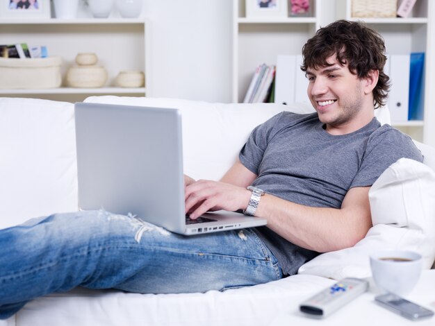 Przystojny młody człowiek relaksujący za pomocą laptopa i leżąc na kanapie
