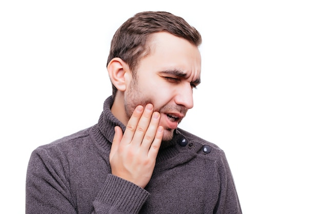 Przystojny młody mężczyzna cierpiący na ból zęba, dotyka policzka, aby powstrzymać ból na białej ścianie