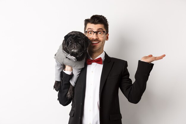 Przystojny młody mężczyzna przedstawiający produkt dla zwierząt domowych pod ręką, trzymający uroczego czarnego psa na ramieniu i uśmiechający się, pokazując coś na białym tle