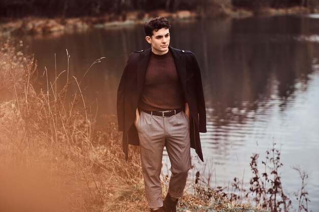 Przystojny młody mężczyzna w płaszczu zakrywającym ramiona, stojąc nad jeziorem w jesiennym lesie.