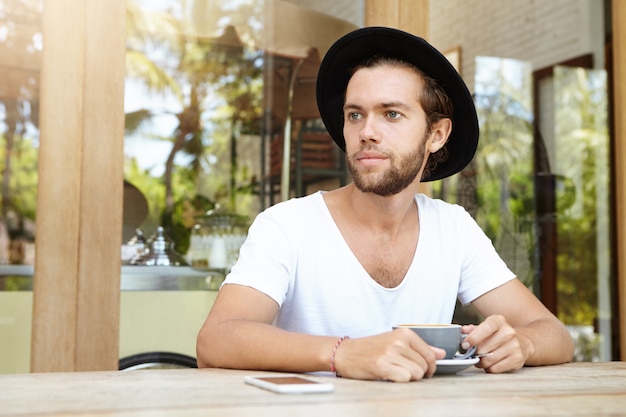 Przystojny młody mężczyzna w stylowym czarnym kapeluszu siedzi przy stole z telefonem komórkowym i kubkiem