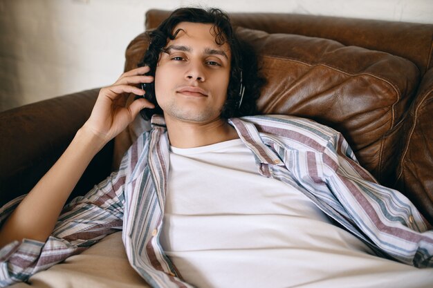 Przystojny, młody mężczyzna z włosiem, leżący na skórzanej kanapie, słuchający nowych utworów online za pośrednictwem usługi strumieniowego przesyłania muzyki przy użyciu słuchawek bezprzewodowych, mając zrelaksowany wygląd.