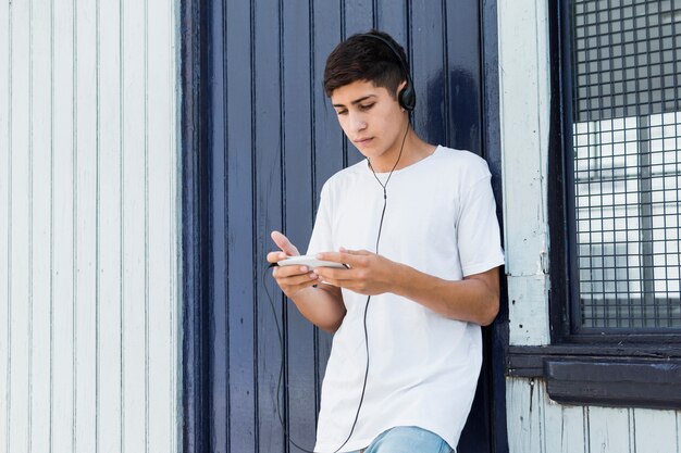 Przystojny nastoletni chłopak opiera na metal ścianie używać smartphone i słuchającą muzykę