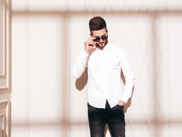 Przystojny pewnie model seksowny stylowy mężczyzna ubrany w białą koszulę i dżinsy Moda hipster mężczyzna pozowanie w pobliżu białej ściany w studio wnętrz w słoneczny dzień cień z okna w okularach przeciwsłonecznych
