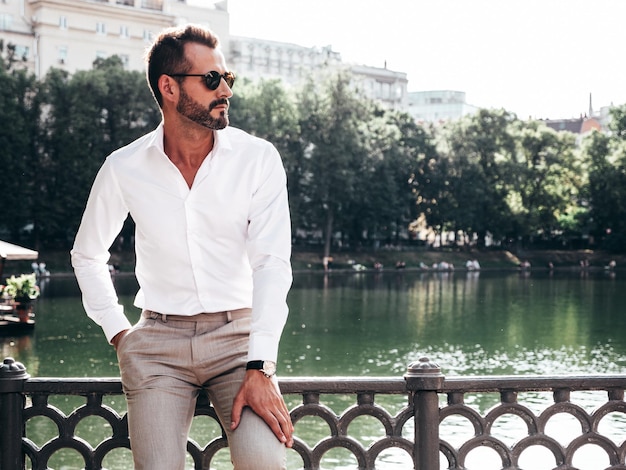 Przystojny pewnie stylowy hipster lamberseksualny modelNowoczesny mężczyzna ubrany w białą koszulę i spodnie Moda mężczyzna pozuje na ulicy w pobliżu nasypu w Europie park miasto o zachodzie słońca w okularach przeciwsłonecznych