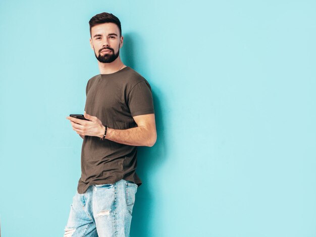 Przystojny pewnie stylowy hipster model lamberseksualnyMężczyzna ubrany w koszulkę i dżinsy Moda mężczyzna na białym tle na niebieskiej ścianie w studioTrzymanie smartfona Patrząc na ekran telefonu Korzystanie z aplikacji