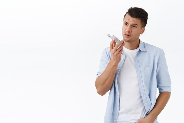 Przystojny, poważny mężczyzna macho w koszuli nagrywa wiadomość głosową lub notatkę w aplikacji na smartfona trzymając telefon komórkowy w pobliżu ust blisko dynamicznej rozmowy z asystentem osoby na białym tle