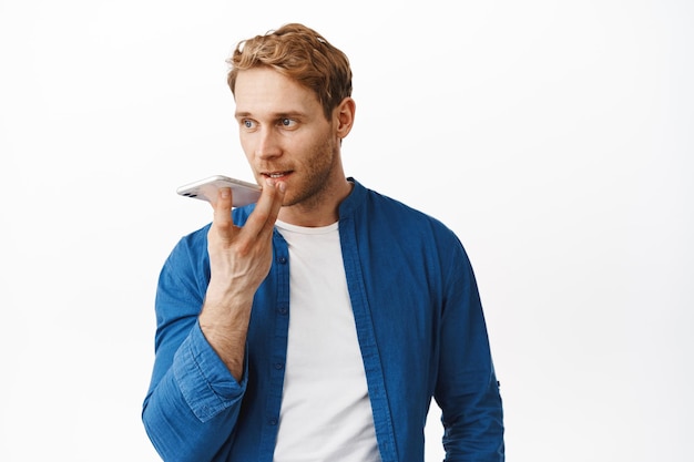 Bezpłatne zdjęcie przystojny rudy mężczyzna nagrywa wiadomość głosową, trzymając smartfon w pobliżu ust, tłumacząc swoje słowa w aplikacji na telefon komórkowy, używając rejestratora aplikacji, stojąc na białym tle