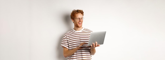 Bezpłatne zdjęcie przystojny rudy mężczyzna pracujący jako niezależny na laptopie uśmiecha się i wygląda na zadowolonego z odrobiny ekranu komputera