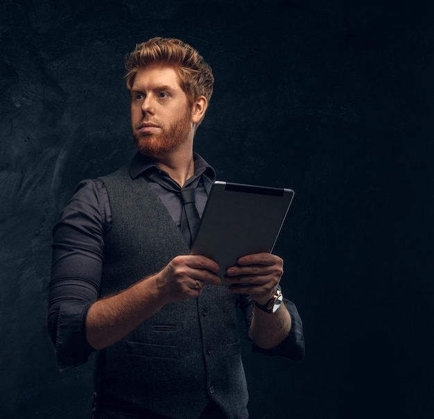 Bezpłatne zdjęcie przystojny rudy mężczyzna w stroju wizytowym, trzymający tablet w studio na tle ciemnej, teksturowanej ściany