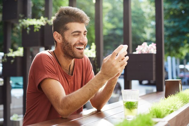 Przystojny szczęśliwy facet siedzi w kawiarni, pije lemoniadę i używa telefonu komórkowego, śmiejąc się z wiadomości tekstowej