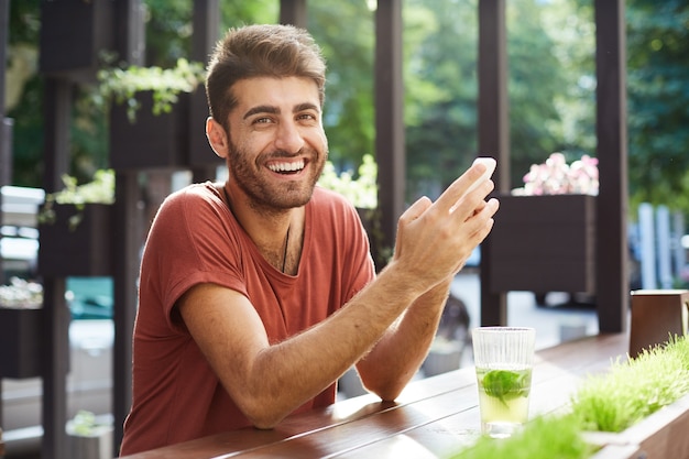 Przystojny szczęśliwy facet siedzi w kawiarni, pije lemoniadę i używa telefonu komórkowego, śmiejąc się z wiadomości tekstowej