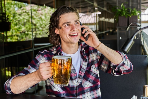 Przystojny uśmiechnięty mężczyzna pije piwo i mówi telefon komórkowy w kawiarni.