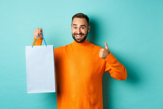 Przystojny uśmiechnięty mężczyzna pokazuje kciuki do góry i torbę na zakupy, polecając sklep, tanding na turkusowej ścianie