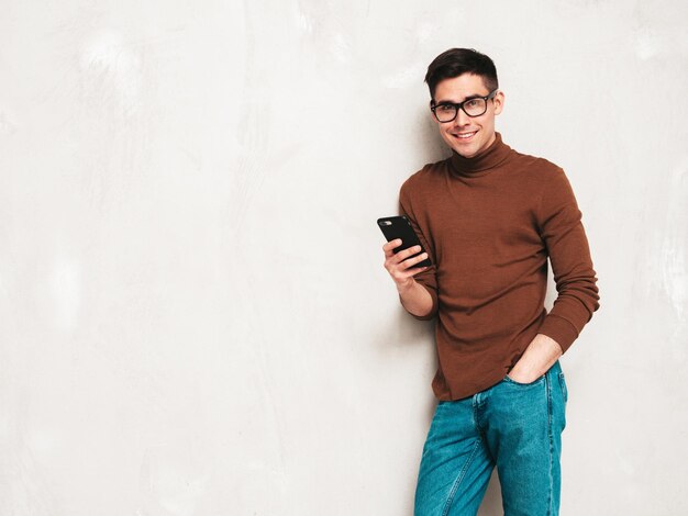 Przystojny uśmiechnięty model Seksowny stylowy mężczyzna ubrany w brązowy sweter z golfem i dżinsy Moda hipster mężczyzna pozuje blisko szarej ściany w studioTrzymanie smartfona Patrząc na ekran telefonu Korzystanie z aplikacji