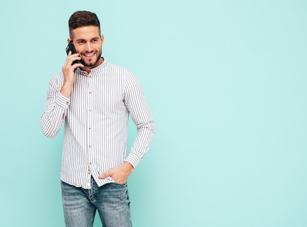 Przystojny uśmiechnięty modelSeksowny stylowy mężczyzna rozmawia przez smartfona Moda hipster mężczyzna pozuje w pobliżu niebieskiej ściany w studio Trzymając telefon Z telefonem komórkowym