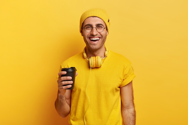Przystojny, wesoły mężczyzna używa słuchawek trzyma kawę na wynos, będąc w dobrym nastroju