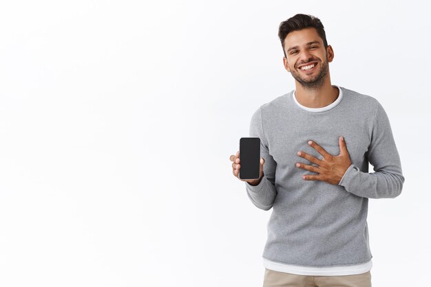 Przystojny zadowolony i schlebiony męski brodaty mężczyzna w szarym swetrze pokazujący swoje wyniki fitness w aplikacji mobilnej dotykający klatki piersiowej wdzięczny lub dumny uśmiechający się radośnie promujący aplikację na smartfonie