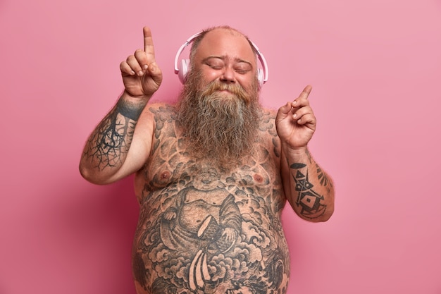 Bezpłatne zdjęcie przystojny, zrelaksowany otyły mężczyzna tańczy do muzyki w słuchawkach, cieszy się każdą piosenką, podnosi ręce i wskazuje palcami, stoi z zamkniętymi oczami, całe ciało pokryte tatuażami.