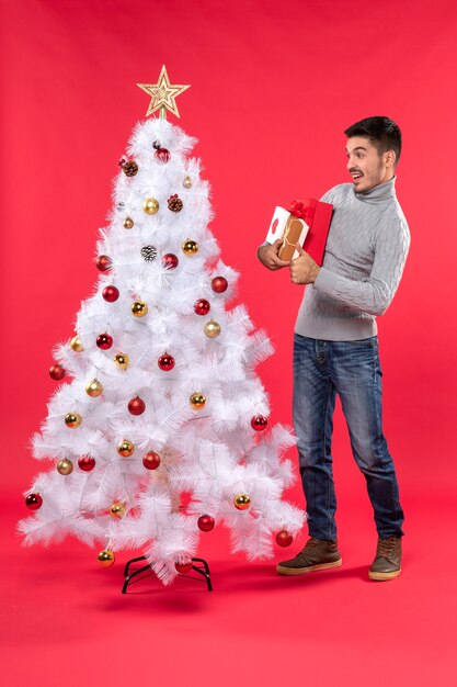 Przystojny zszokowany młody człowiek stojący w pobliżu udekorowanego białego drzewa nowego roku i trzymając swoje prezenty
