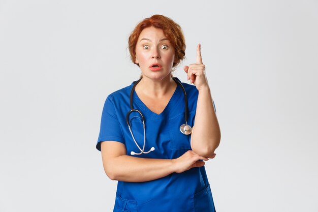 Przytłoczona rudowłosa lekarka, pielęgniarka w średnim wieku w fartuchu mają sugestie, mówiąc pomysł lub plan, unosząc palec wskazujący gestem eureki z zatroskaną miną, stojąc