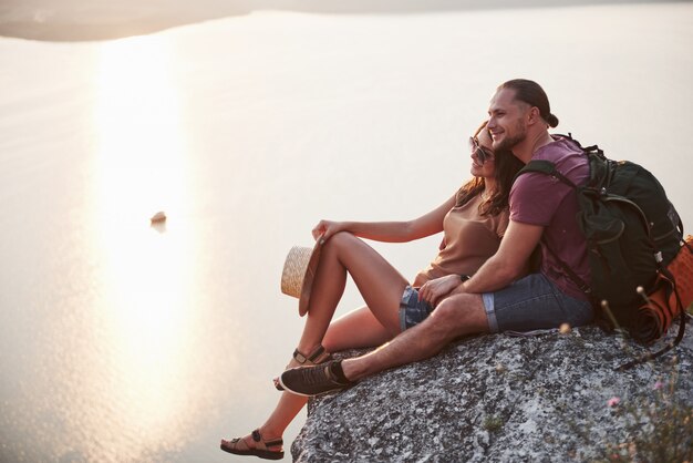 Przytulanie para z plecakiem siedząc na szczycie skały, ciesząc się widokiem wybrzeża rzeki lub jeziora.