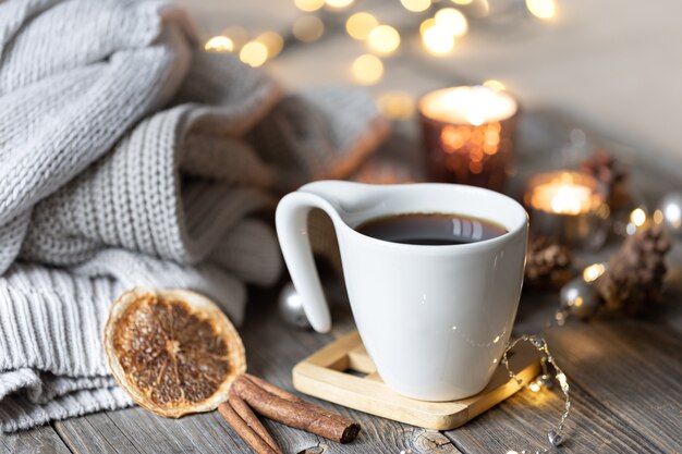 Przytulna domowa zimowa kompozycja z filiżanką herbaty na rozmytym tle z płonącymi świecami i światłami bokeh i dzianinowymi elementami.