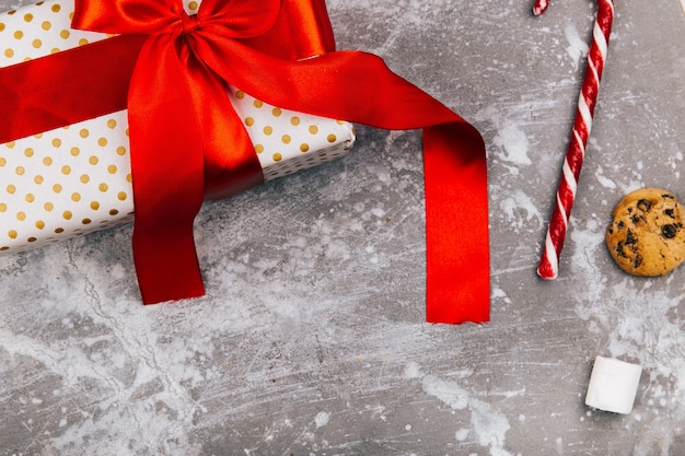 Pudełko z czerwoną wstążką leży na szarej podłodze z świątecznymi ciasteczkami, pierniczkami i czerwonymi białymi cukierkami