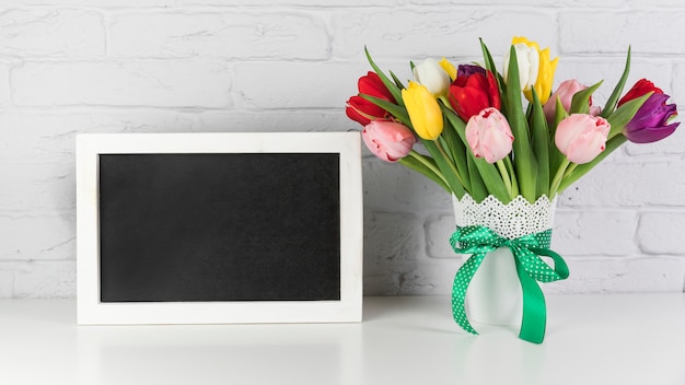 Bezpłatne zdjęcie pusta czarna rama z tulipan wazą na biurku przeciw białemu ściana z cegieł