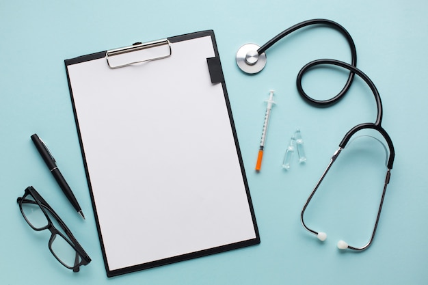 Pusty biały papier w schowku w pobliżu stetoskopu; iniekcja; długopis i okulary na niebieskim biurku