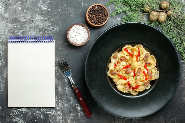 Pyszne conchiglie z warzywami i zieleniną na talerzu i nożem oraz różnymi przyprawami obok notatnika na szarym tle