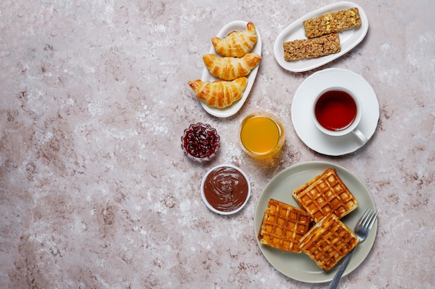 Pyszne śniadanie z kawą, sokiem pomarańczowym, goframi, rogalikami, dżemem, pastą orzechową na świetle, widok z góry