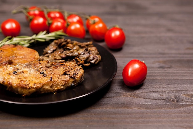 Bezpłatne zdjęcie pyszny stek wieprzowy w czarnym talerzu z grillowanymi pieczarkami, pomidorkami cherry oregano i pastą z fasoli na drewnianym stole