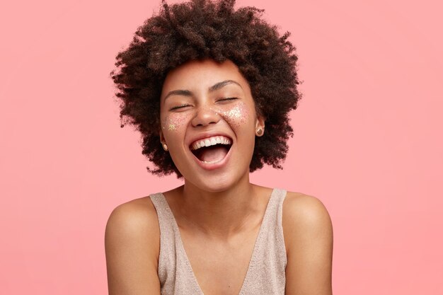 Radosna Afroamerykanka o ciemnej karnacji, radośnie się śmieje, szeroko otwiera usta, ma iskierki na policzkach, zamyka oczy, ma kręcone włosy, odizolowane na różowej ścianie. Koncepcja ludzi i szczęścia