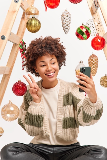 Radosna Afroamerykanka robi selfie pokazuje gest pokoju trzyma telefon komórkowy nosi swobodny sweter ma radosne pozy