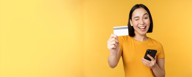 Radosna Azjatycka dziewczyna uśmiecha się pokazując kartę kredytową i smartfona, polecając bankowość komórkową stojącą na żółtym tle