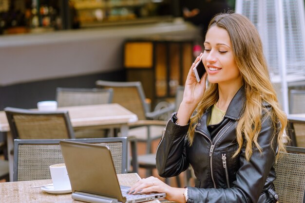 Radosna blondynki kobieta siedzi w kawiarni na zewnątrz rozmawia przez telefon i korzysta z laptopa