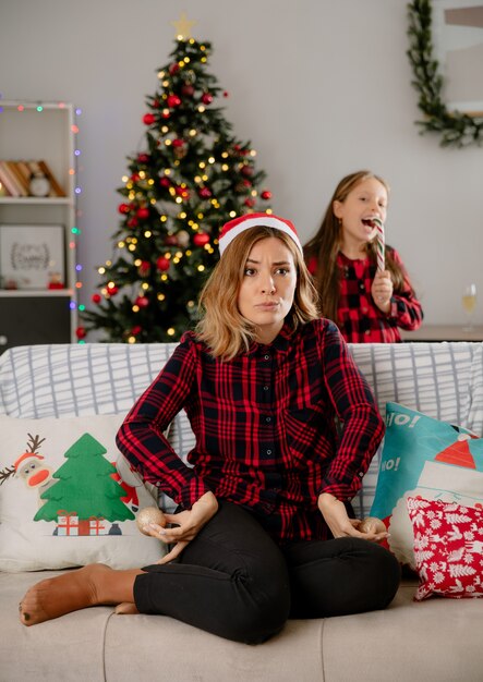Radosna córka jedząca cukierkową laskę i zła matka trzyma szklane kulki, siedząc na kanapie i ciesząc się świątecznymi chwilami w domu