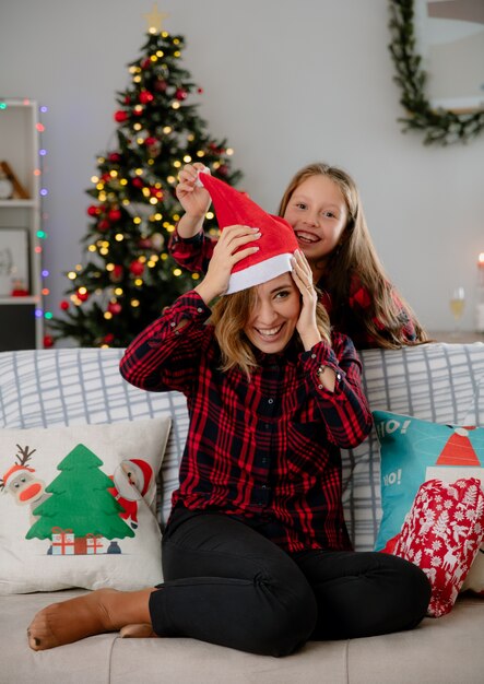 Radosna córka wkłada czapkę Mikołaja na głowę matki siedzącej na kanapie i ciesząc się świątecznymi chwilami w domu