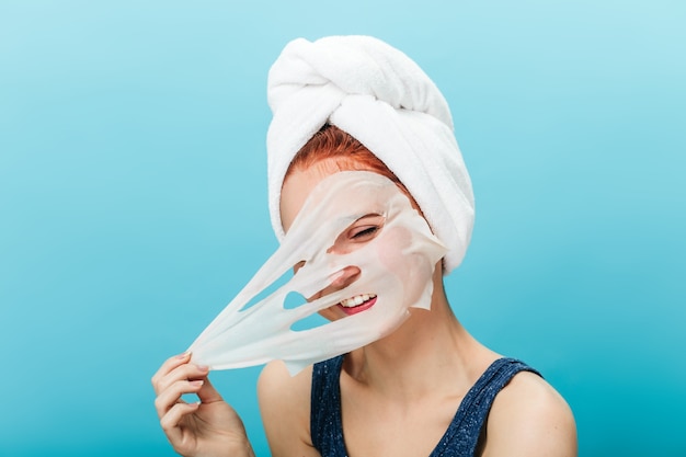 Bezpłatne zdjęcie radosna dziewczynka kaukaski zdejmująca maskę. studio strzał beztroski młoda kobieta robi zabiegi pielęgnacji skóry na niebieskim tle.