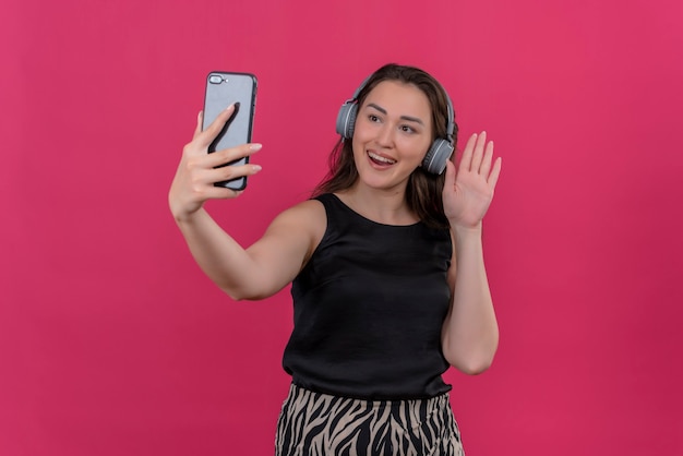 Radosna kobieta ubrana w czarny podkoszulek w słuchawkach rozmawia przez telefon na różowej ścianie