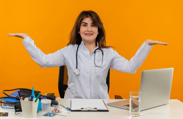 Radosna kobieta w średnim wieku ubrana w szlafrok medyczny i stetoskop siedząca przy biurku ze schowkiem na narzędzia medyczne i laptopem, pokazująca puste ręce na białym tle