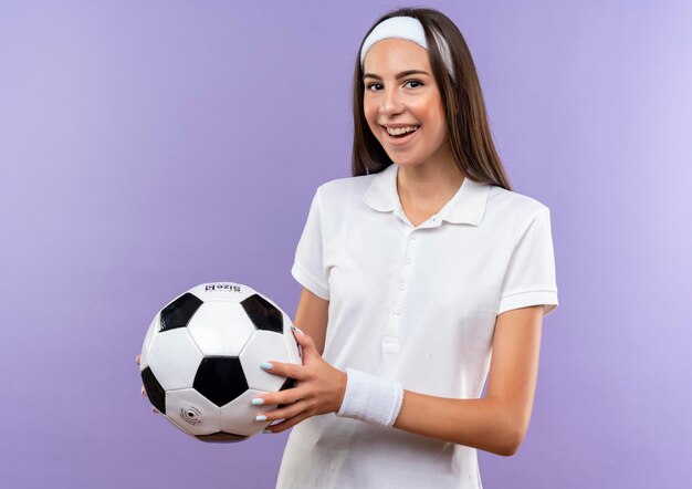 Radosna ładna sportowa dziewczyna nosząca opaskę na głowę i nadgarstek trzymająca piłkę nożną odizolowaną na fioletowej ścianie