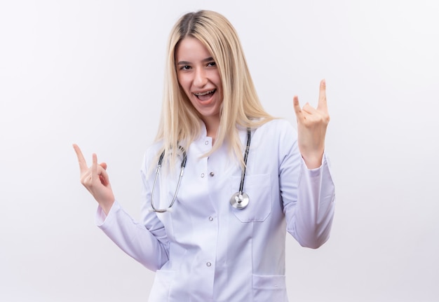 Radosna lekarka młoda blondynka ubrana w stetoskop i fartuch medyczny w ortezie dentystycznej pokazująca gest kozła obiema rękami na odizolowanej białej ścianie