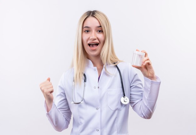 Radosna lekarka młoda blondynka ubrana w stetoskop i fartuch medyczny w ortezie dentystycznej trzymająca pustą puszkę pokazująca gest tak na odizolowanej białej ścianie